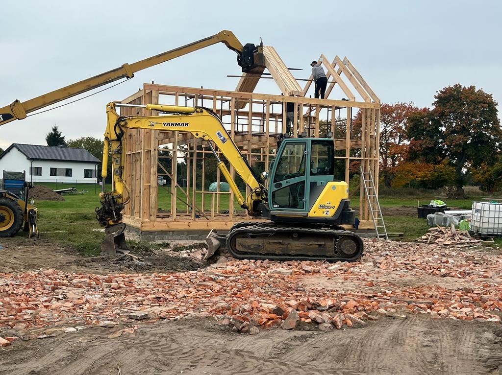 Żółta minikoparka Yanmar z podnoszonym ramieniem pomaga w budowie drewnianej konstrukcji domu, robotnicy montują więźbę dachową, w tle biały budynek i drzewa, na pierwszym planie gruz ceglany.