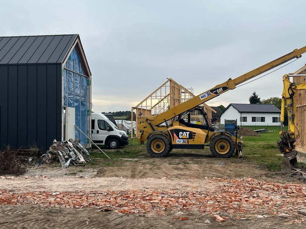 Żółta ładowarka teleskopowa CAT na placu budowy z widocznymi elementami konstrukcji domu w tle, gruzem i stertą cegieł na pierwszym planie.