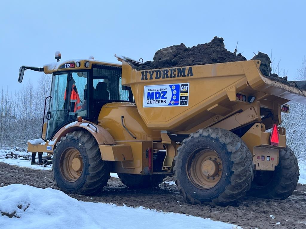 Żółta wywrotka Hydrema z ładunkiem ziemi na śniegu, widoczne logo firmy MDZ Zuterek i CAT, operator w kabinie w pomarańczowej kamizelce odblaskowej.