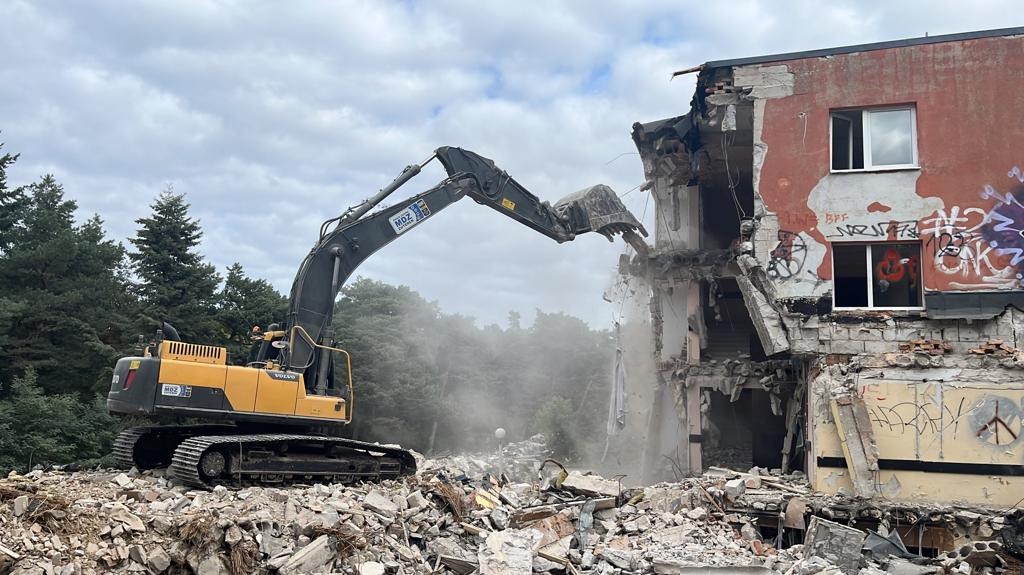 Żółto-czarna koparka gąsienicowa Volvo HB68 kruszy fragment budynku z graffiti na tle drzew i pochmurnego nieba.