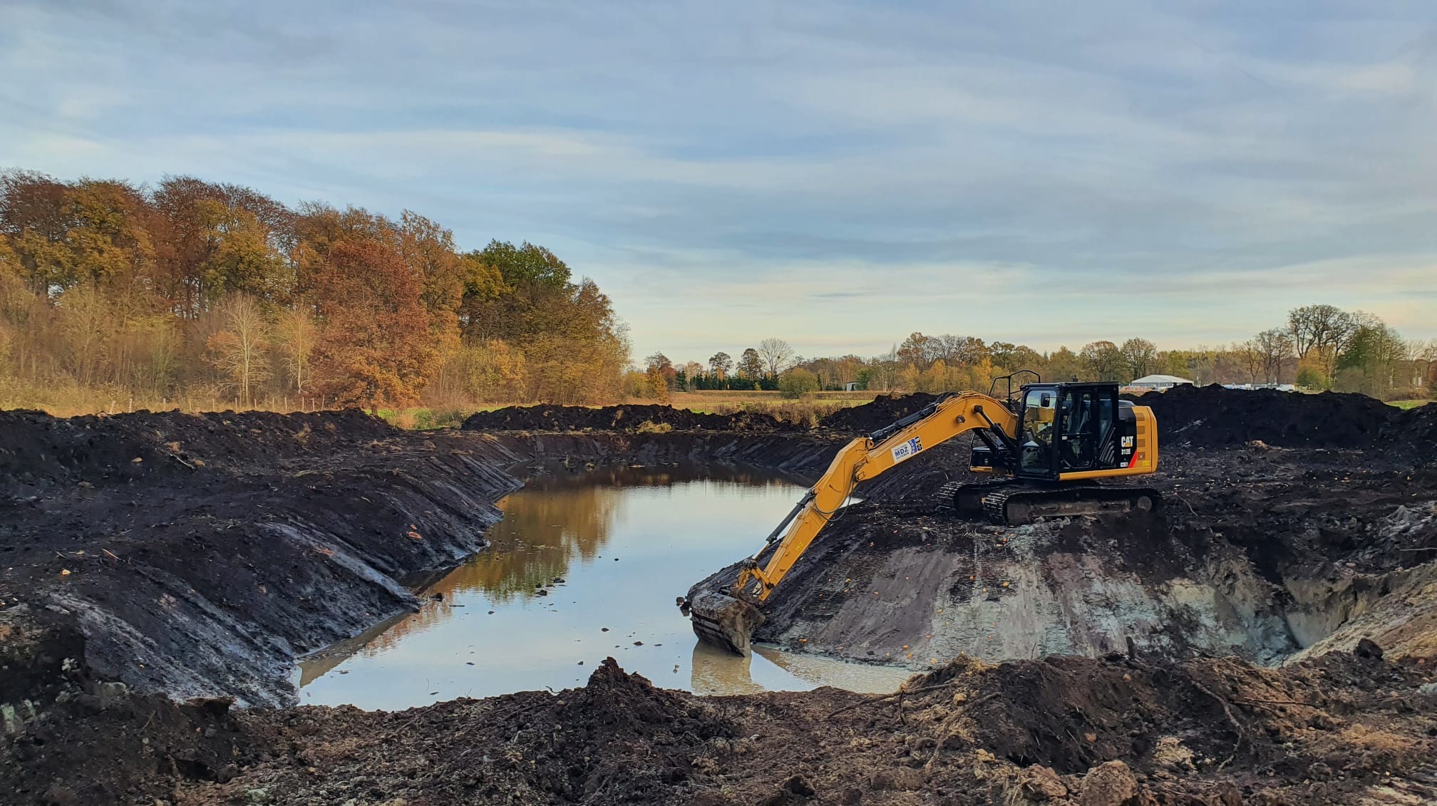 Żółta koparka CAT 312E na skraju wykopu wypełnionego wodą, z widocznymi śladami pracy i świeżo wykopaną ziemią, jesienne drzewa w tle pod pochmurnym niebem.