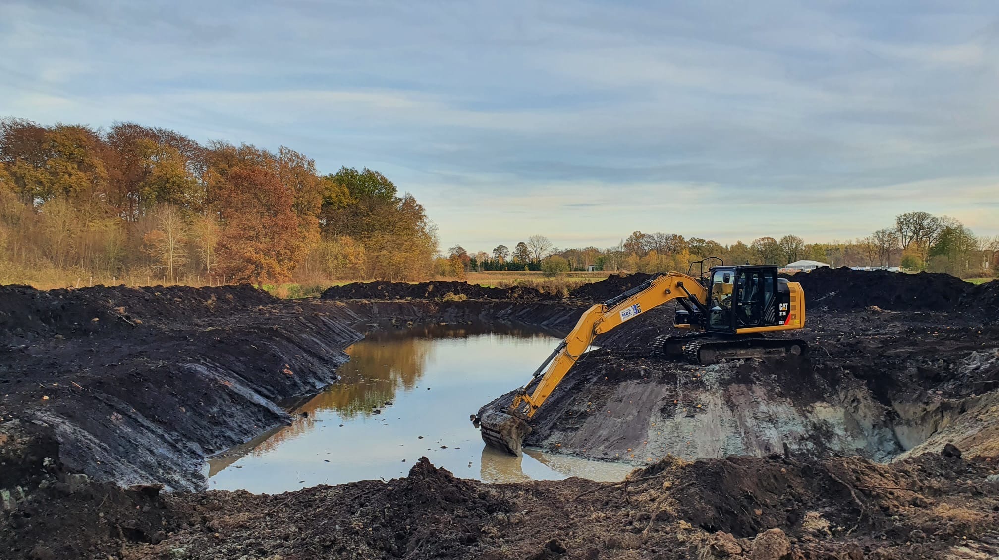 Żółta koparka CAT 312E na skraju wykopu wypełnionego wodą, otoczonego brzegami z ciemnej ziemi, w tle drzewa z jesiennymi liśćmi i pochmurne niebo.