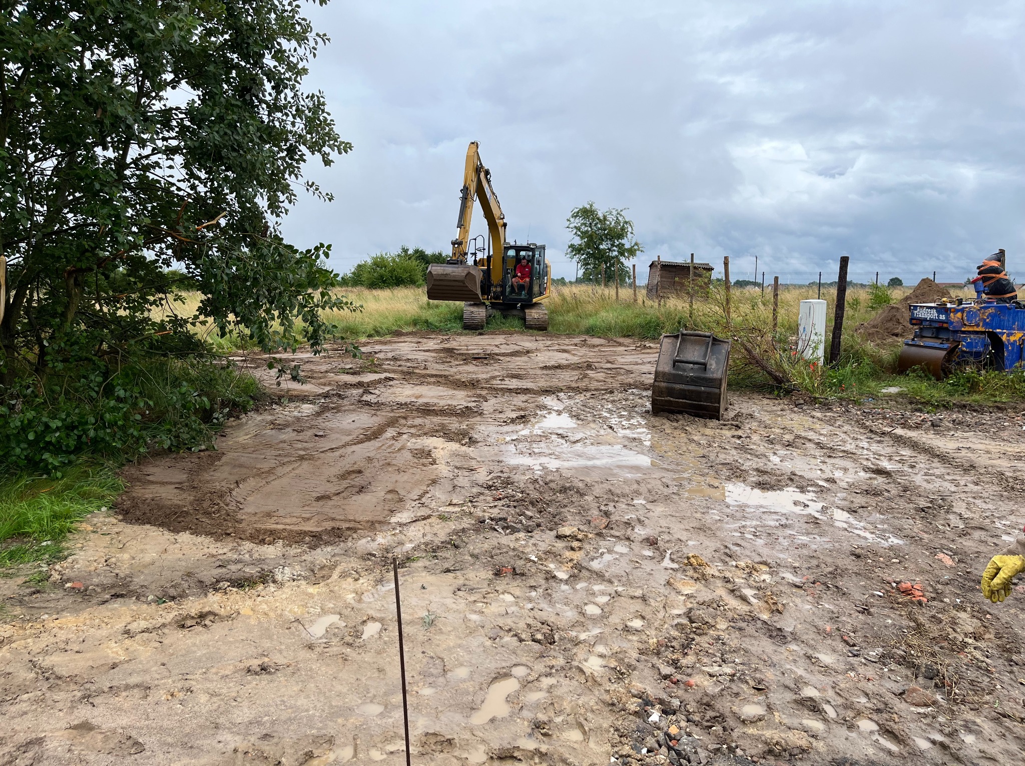 Żółta koparka na rozmokniętym terenie budowy, widoczny operator w kabinie, w tle zarośla i pochmurne niebo, na pierwszym planie błoto i ślady opon, obok walec drogowy i kontener.