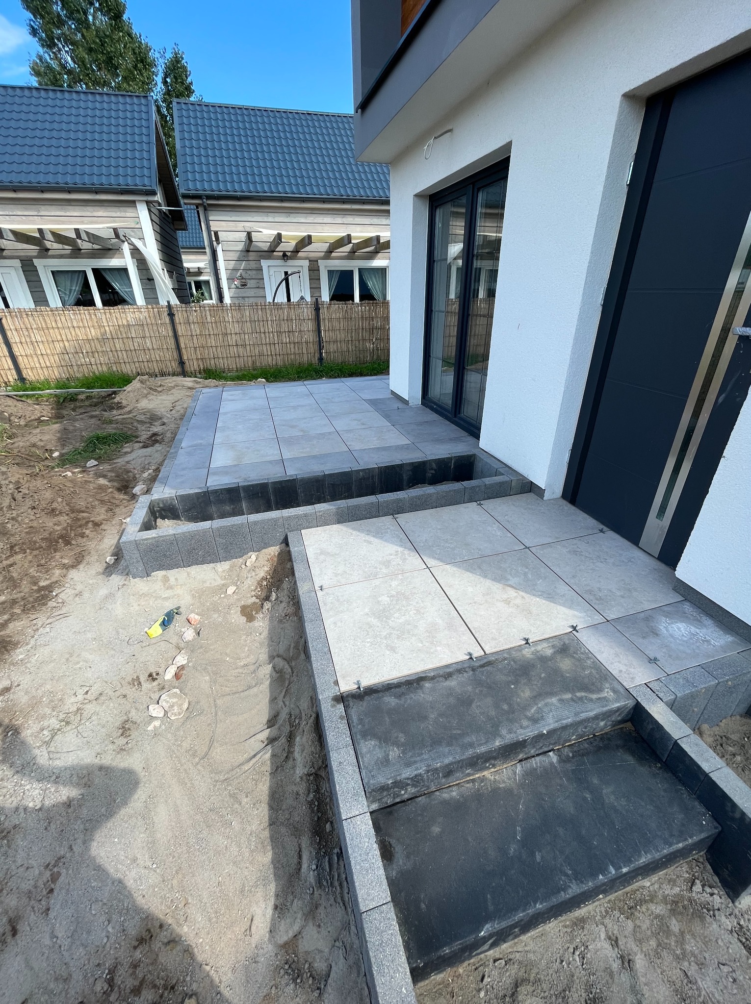 Układanie szarej kostki brukowej na tarasie przed nowoczesnym domem z czarnymi drzwiami, widoczne plastikowe krzyżyki dystansowe i fragmenty podłoża w trakcie prac.