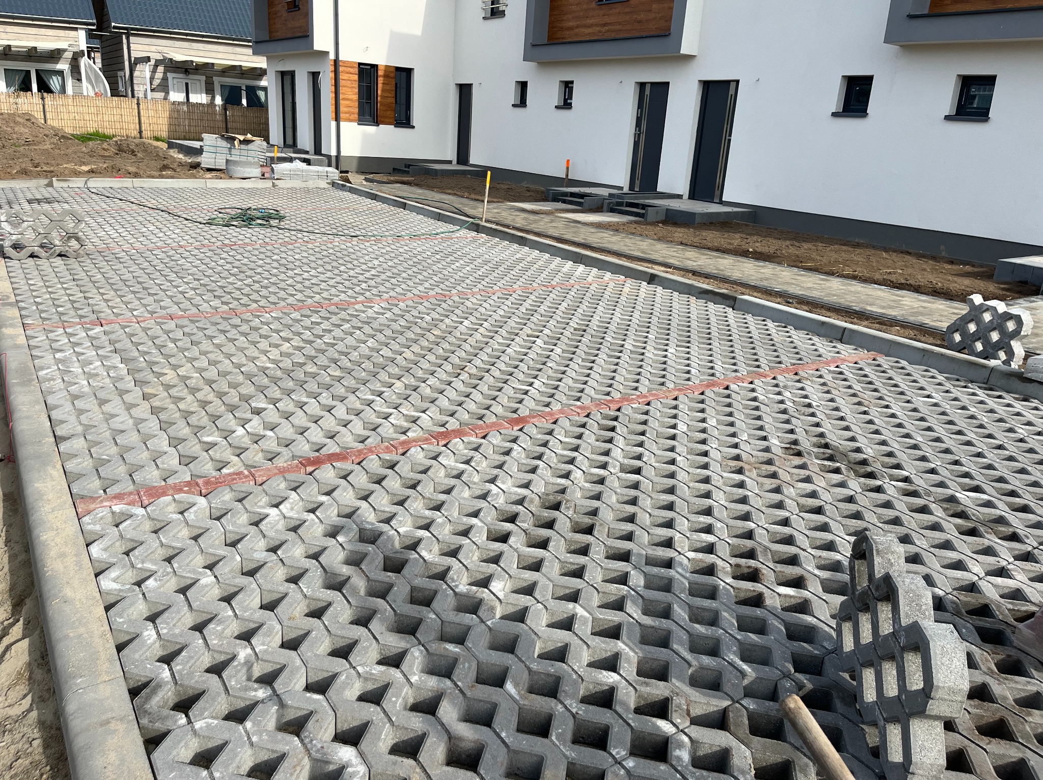 Układanie ażurowych płyt betonowych na przygotowanym podłożu, widoczne świeżo ułożone obrzeża i fragmenty niezakończonej powierzchni, w tle budynki mieszkalne.