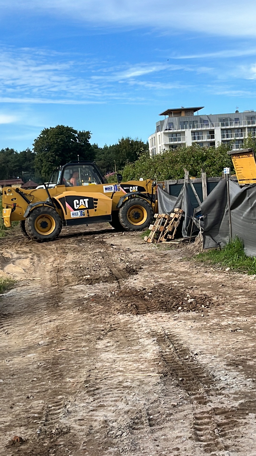 Żółta ładowarka teleskopowa marki CAT na placu budowy z widocznymi śladami opon na ziemi, w tle budynek mieszkalny i drzewa, pod błękitnym niebem.