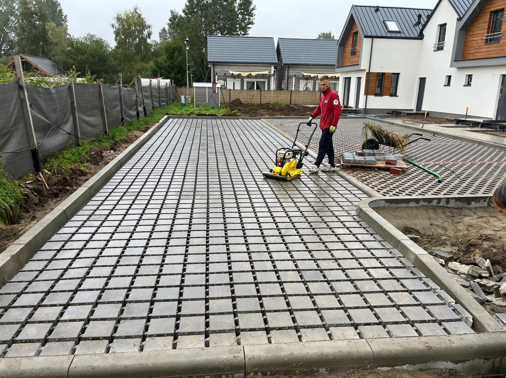 Pracownik zagęszcza podłoże pod ażurowe płyty betonowe na podjeździe nowo wybudowanego domu jednorodzinnego przy użyciu zagęszczarki wibracyjnej.
