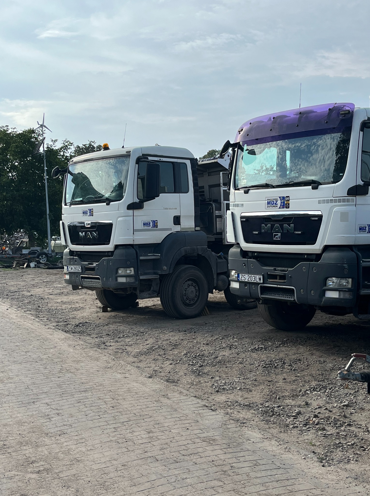 Dwa białe samochody ciężarowe MAN zaparkowane na żwirowym placu, jeden z fioletową osłoną przeciwsłoneczną, w tle widoczna turbina wiatrowa.