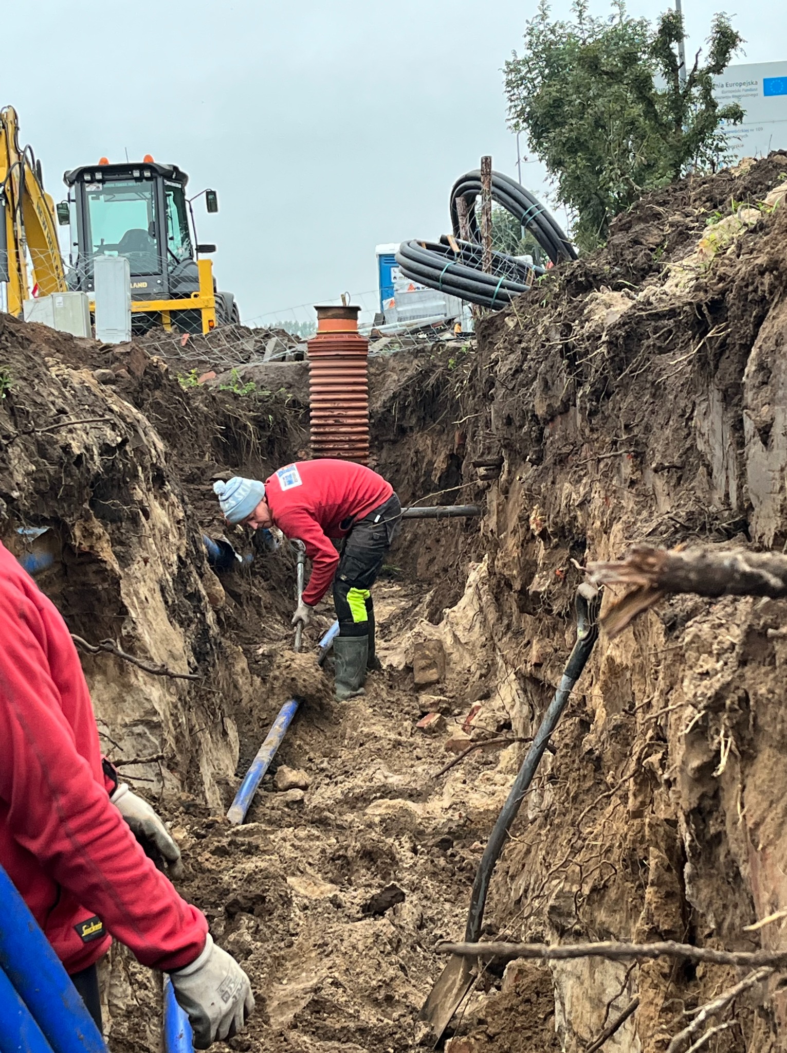 Pracownik w czerwonej bluzie i czapce pracuje w głębokim wykopie, układając niebieskie rury, w tle widoczna rura kanalizacyjna z tworzywa sztucznego i żółta koparka.