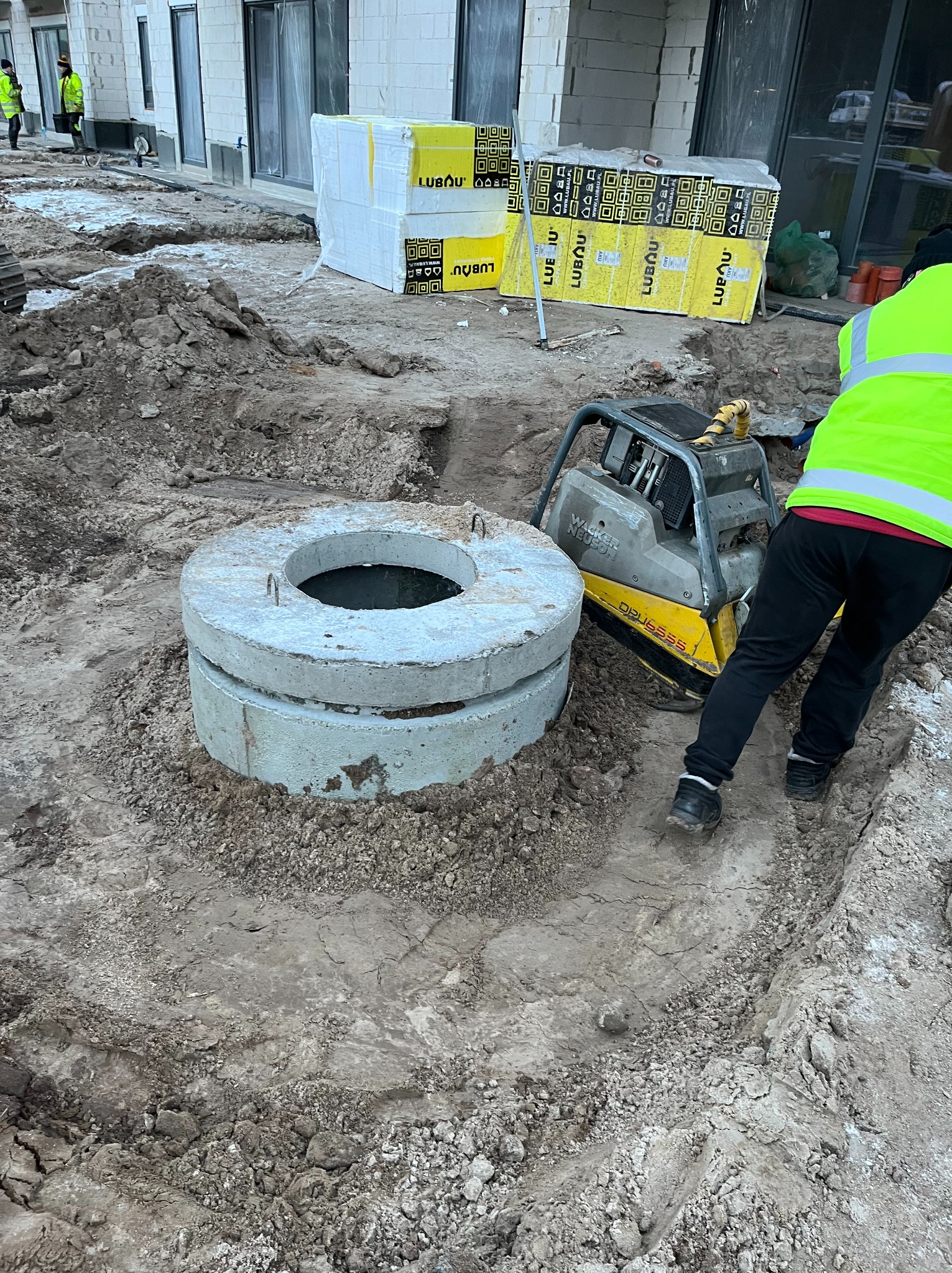 Dwa betonowe kręgi studni na wykopie, ubijarka Wacker Neuson obok, pracownik w kamizelce odblaskowej, materiały budowlane Lubau w tle.