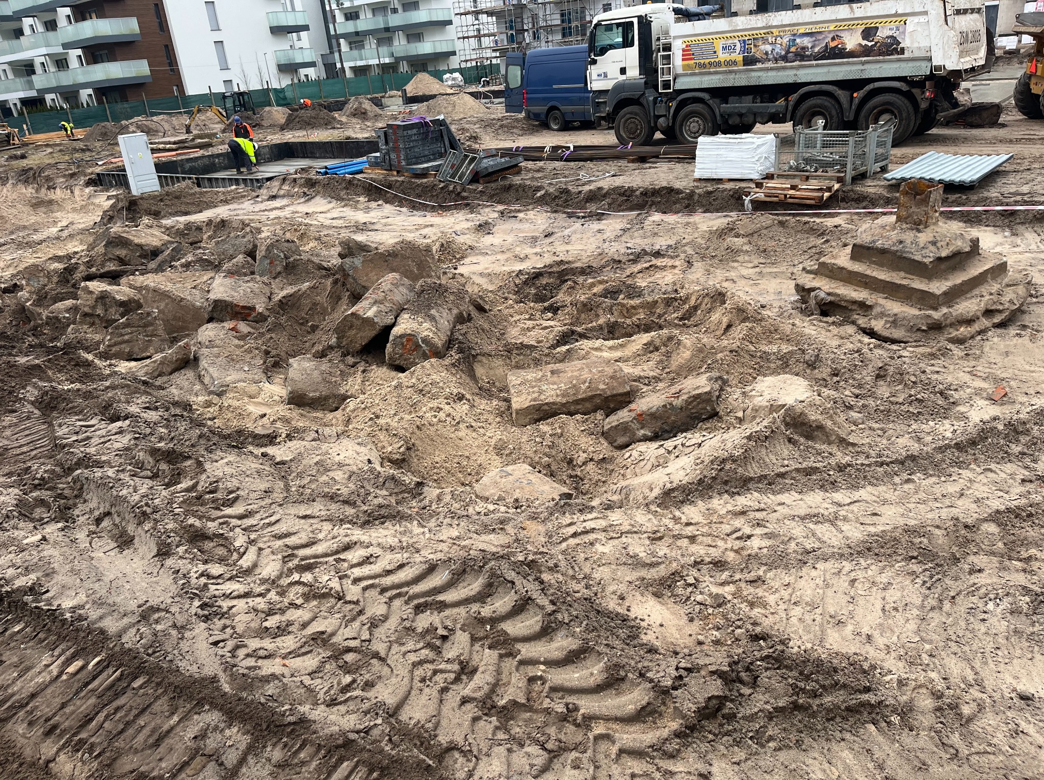 Teren budowy z wykopem, gruzem i śladami opon ciężkiego sprzętu, w tle budynki mieszkalne i ciężarówka z reklamą robót ziemnych.