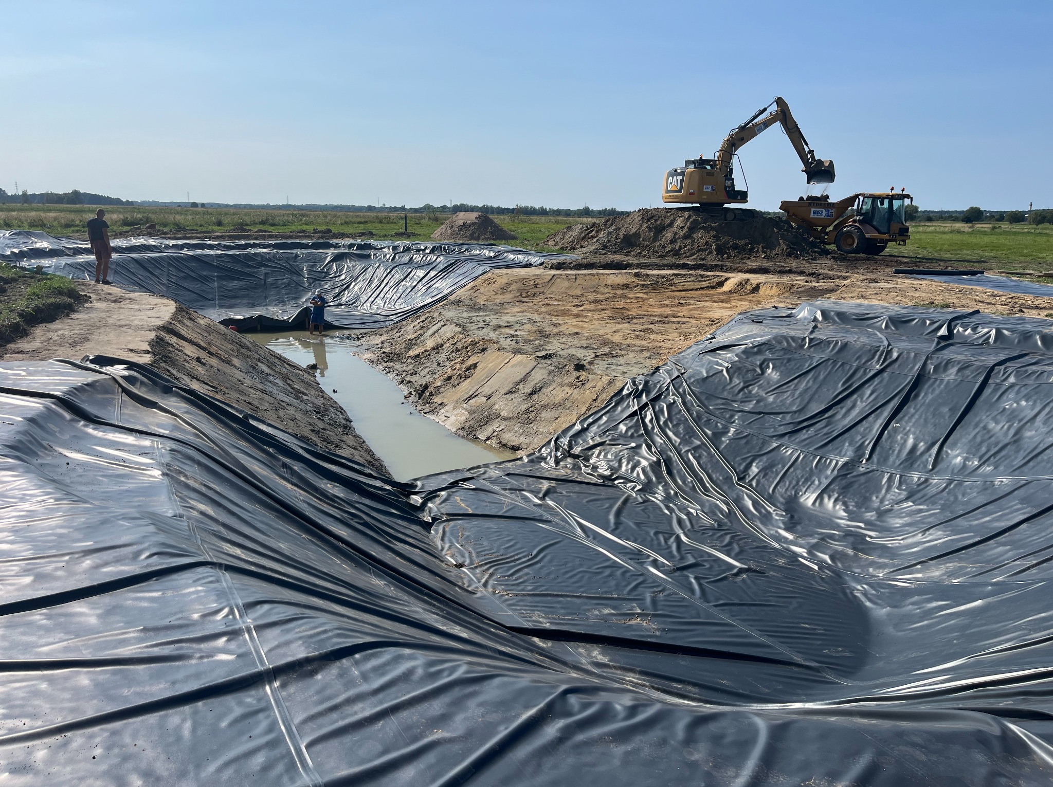 Wyłożony czarną folią wykop pod zbiornik wodny na polu, z pracującą koparką CAT i dwiema osobami w tle.
