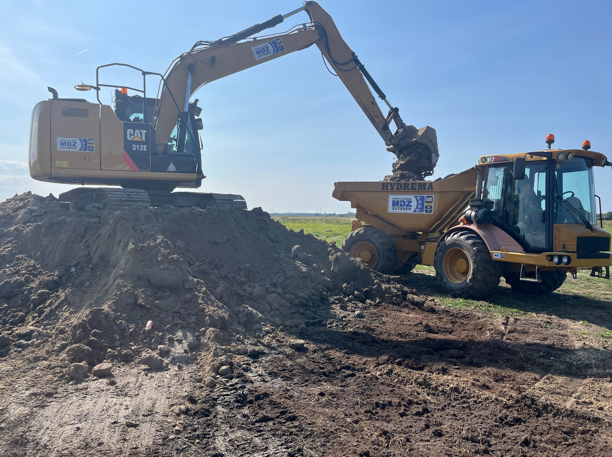 Żółta koparka CAT 312E nabiera ziemię z pryzmy i wrzuca ją do wywrotki Hydrema na tle pola i błękitnego nieba.