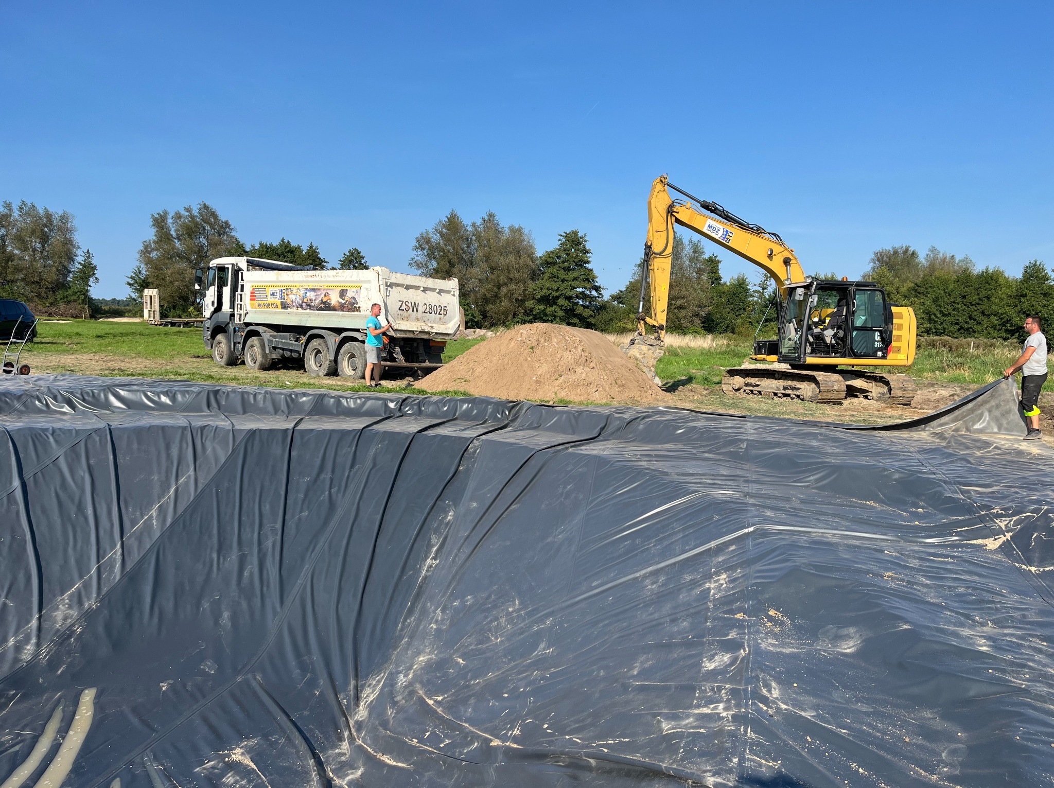 Wykop wyłożony czarną folią, obok koparka i wywrotka z ziemią, dwóch mężczyzn przy pracy, błękitne niebo w tle.