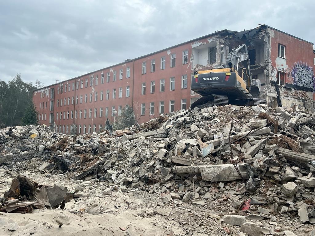 Żółto-czarna koparka Volvo na szczycie sterty gruzu po rozbiórce budynku, z widoczną częścią wielopiętrowego budynku w tle i pochmurnym niebem.