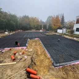 DonBud - Przygotowany teren pod fundamenty z rozłożoną siatką zbrojeniową na izolacji termicznej, widoczne rury kanalizacyjne i wykop w piaszczystej ziemi, drzewa w tle spowite mgłą.