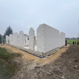 DonBud - Wznoszenie ścian parterowego domu z białych bloczków na fundamencie, widok z zewnątrz w pochmurny dzień.