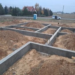 DonBud - Zalane betonem szalunki fundamentowe z wystającymi prętami zbrojeniowymi, przygotowane do dalszych prac budowlanych, na tle krajobrazu z domem i przenośną toaletą.