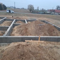 DonBud - Wylane betonowe fundamenty pod budynek z wystającymi prętami zbrojeniowymi, widok na rozkopany teren i samochód dostawczy w tle.