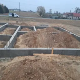 Wylane betonowe fundamenty pod budynek z wystającymi prętami zbrojeniowymi, widok na rozkopany teren i samochód dostawczy w tle.