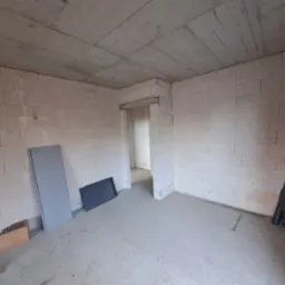 Surowy stan wnętrza budynku w trakcie budowy: ściany z betonu komórkowego, surowy betonowy strop, otwór drzwiowy bez ościeżnicy, fragmenty materiałów budowlanych na podłodze.