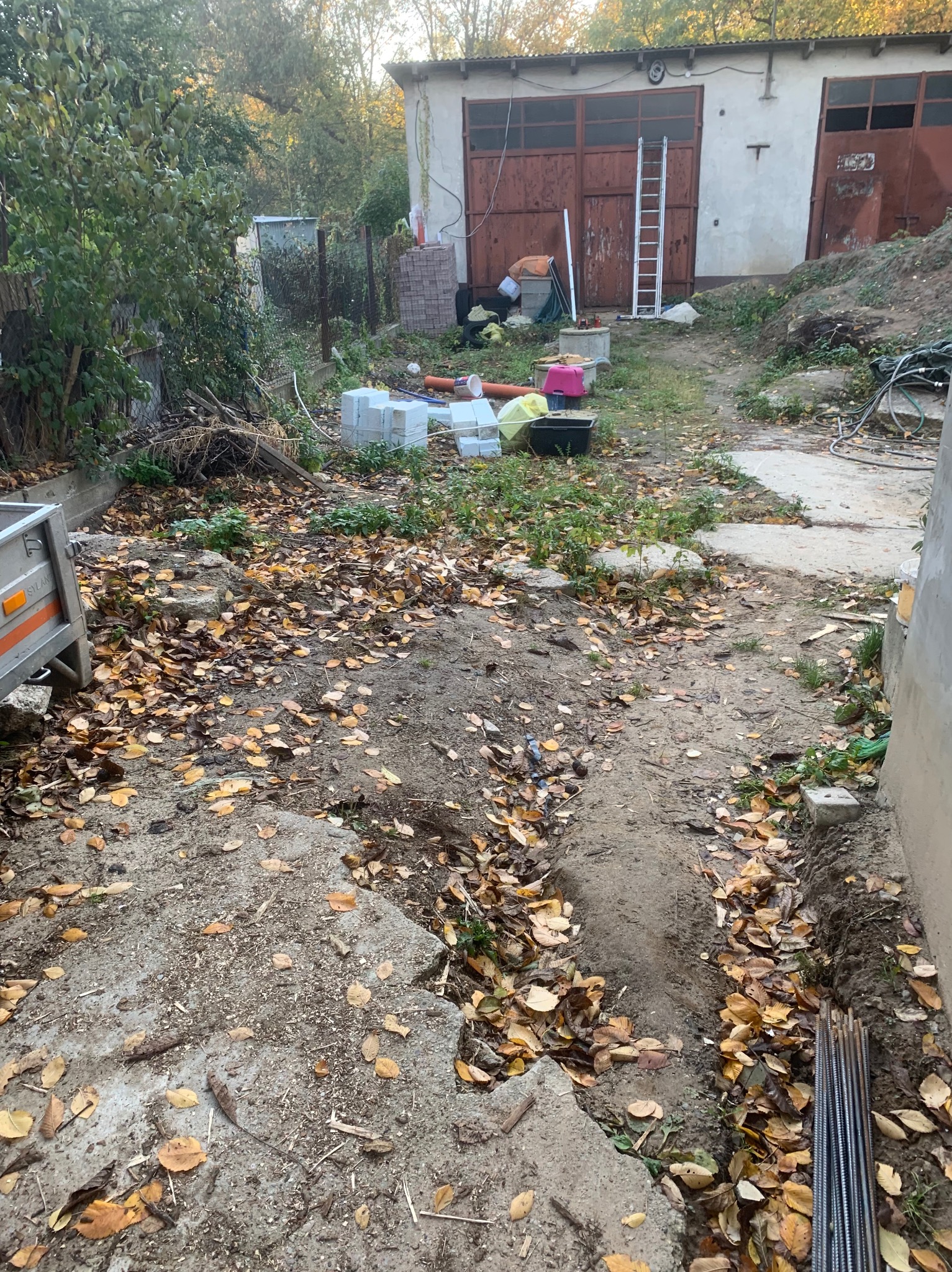 Przygotowanie terenu pod fundamenty: wykop, betonowe elementy, zbrojenie, liście i budynek gospodarczy w tle.