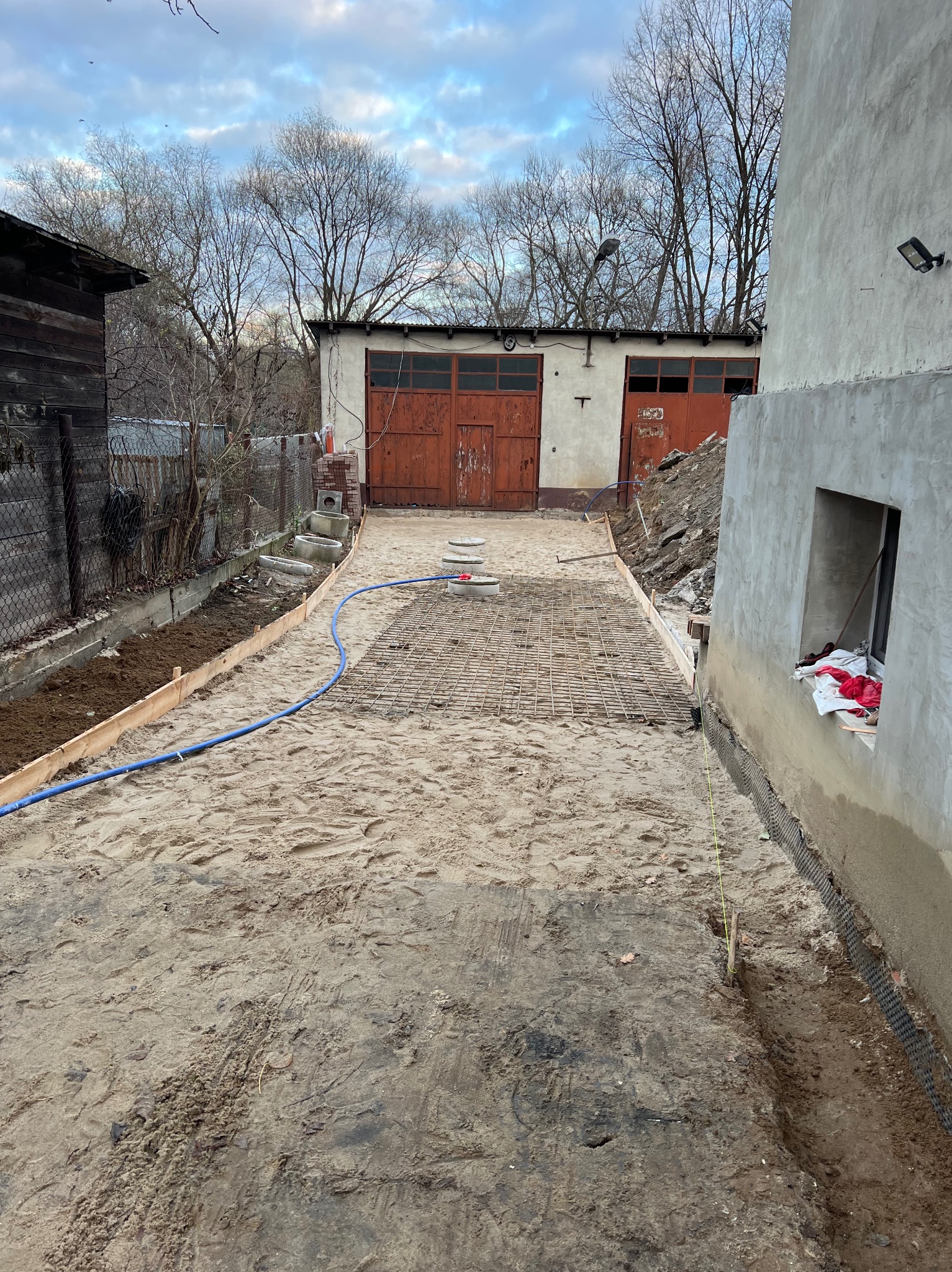 Przygotowanie terenu pod wylewkę: szalunki, siatka zbrojeniowa, piasek, widok na garaż z czerwonymi bramami w tle.