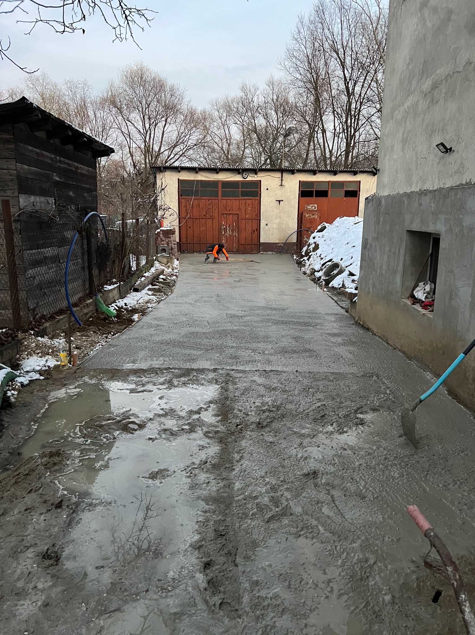 Świeżo wylana betonowa droga do garażu, pracownik w pomarańczowej kurtce wyrównuje powierzchnię, obok leżą narzędzia i resztki śniegu.