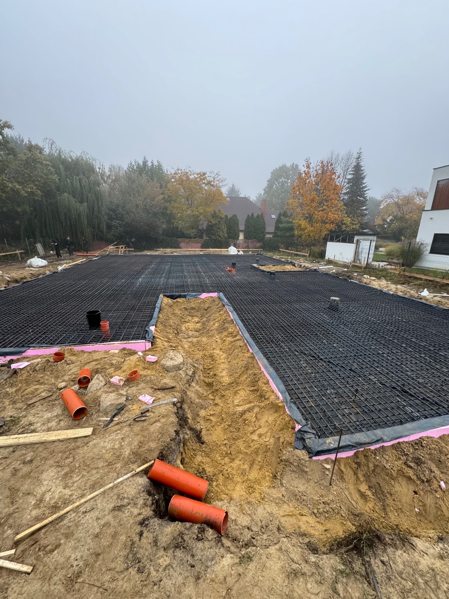 Przygotowany teren pod fundamenty z rozłożoną siatką zbrojeniową na izolacji termicznej, widoczne rury kanalizacyjne i wykop w piaszczystej ziemi, drzewa w tle spowite mgłą.