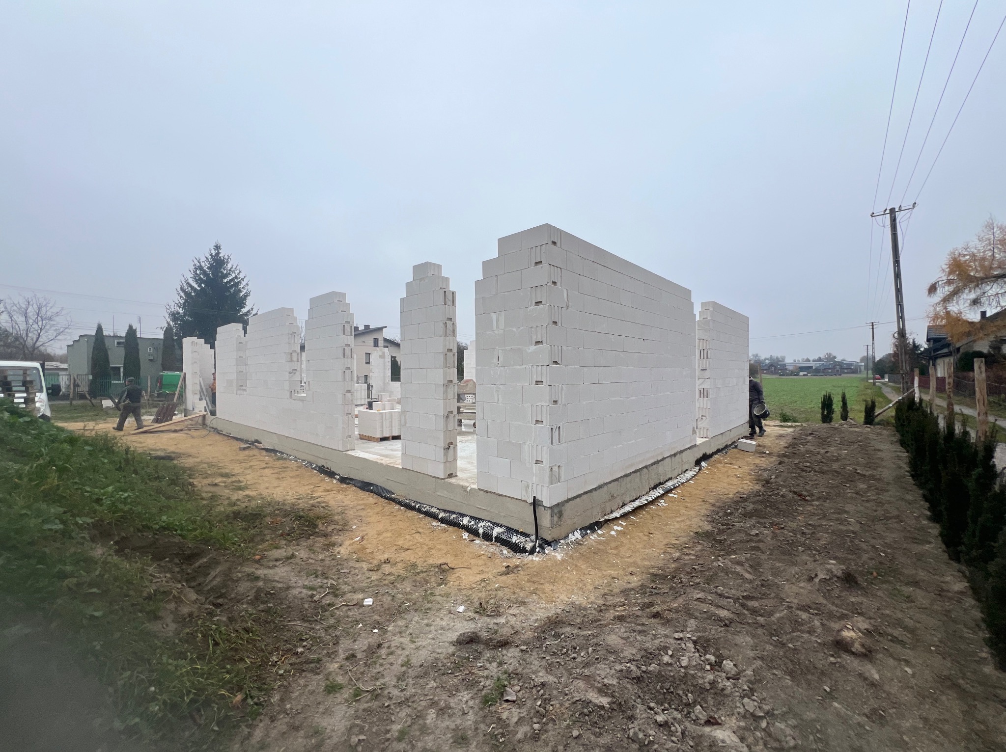 Wznoszenie ścian parterowego domu z białych bloczków na fundamencie, widok z zewnątrz w pochmurny dzień.