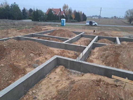 Zalane betonem szalunki fundamentowe z wystającymi prętami zbrojeniowymi, przygotowane do dalszych prac budowlanych, na tle krajobrazu z domem i przenośną toaletą.