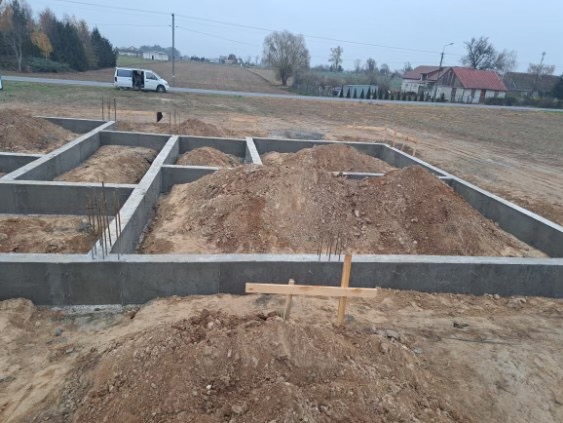 Wylane betonowe fundamenty pod budynek z wystającymi prętami zbrojeniowymi, widok na rozkopany teren i samochód dostawczy w tle.