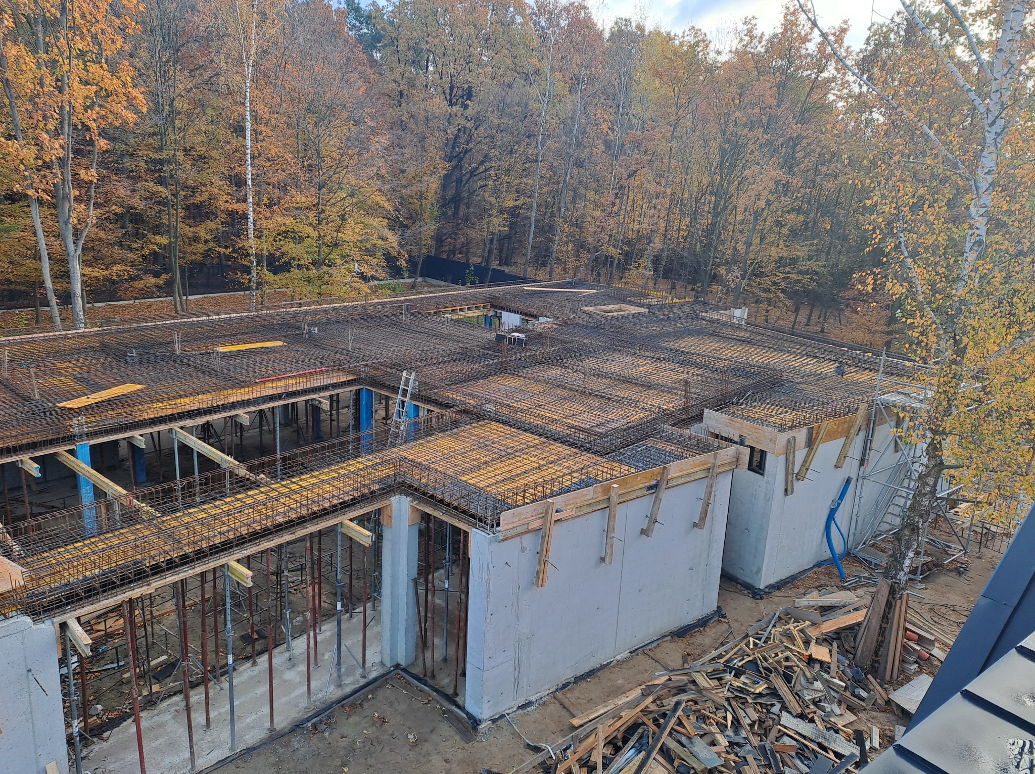 Wysoki kąt widzenia na plac budowy w jesiennej scenerii, widoczne zbrojenie stropu, betonowe ściany oraz tymczasowe podpory, w tle las z drzewami o żółtych liściach.