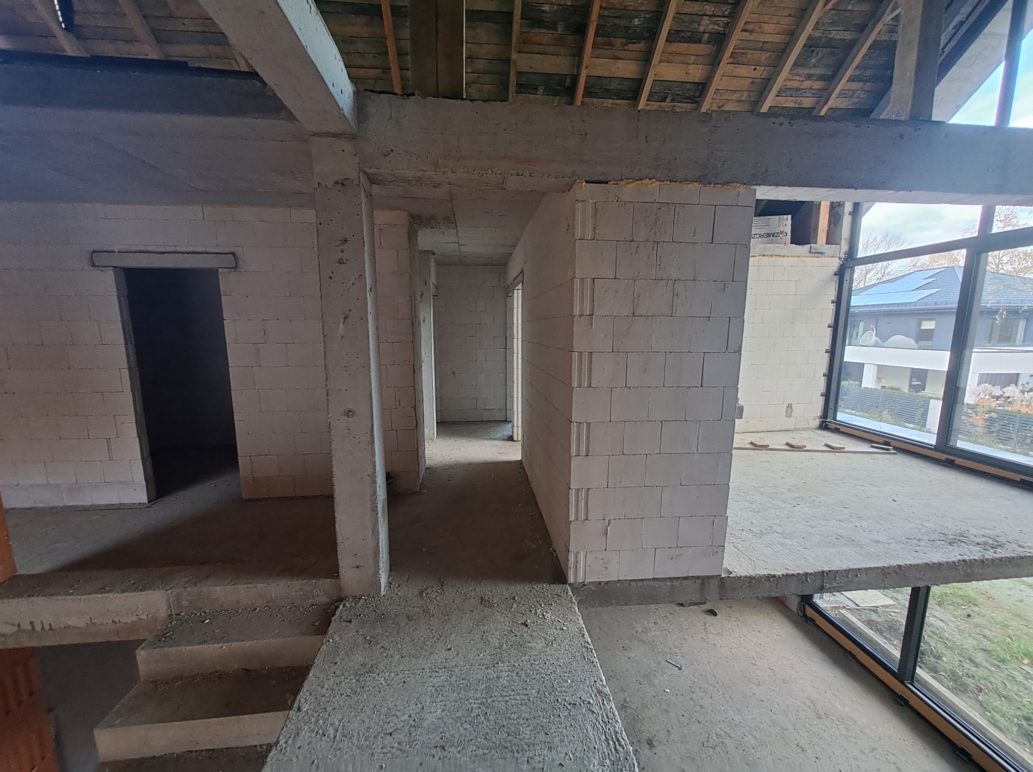 Surowy stan wnętrza domu w trakcie budowy z widocznymi ścianami z białego bloczka, betonowymi schodami i drewnianym stropem, duże okno z widokiem na zewnątrz.