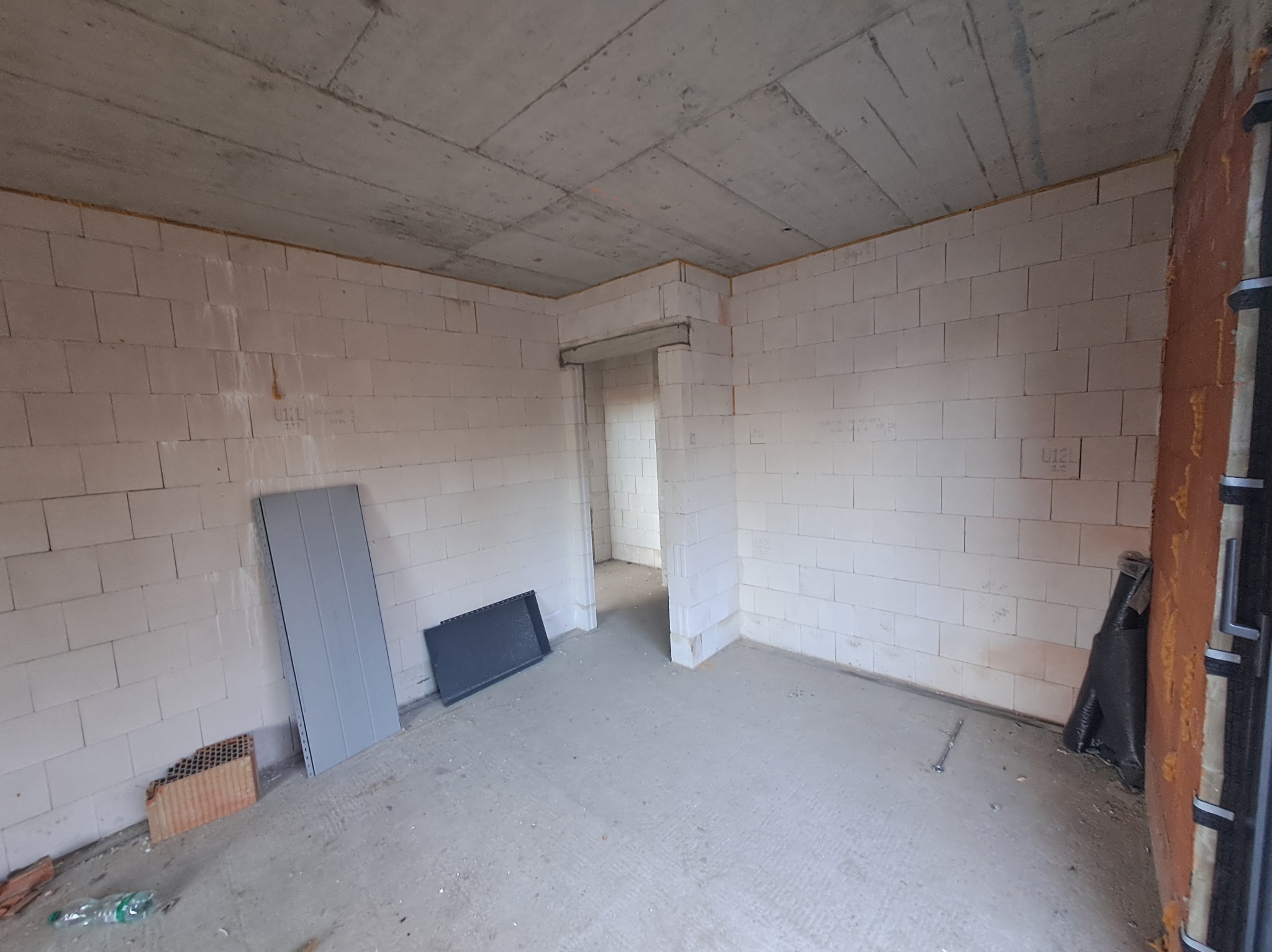 Surowy stan wnętrza budynku w trakcie budowy: ściany z betonu komórkowego, surowy betonowy strop, otwór drzwiowy bez ościeżnicy, fragmenty materiałów budowlanych na podłodze.