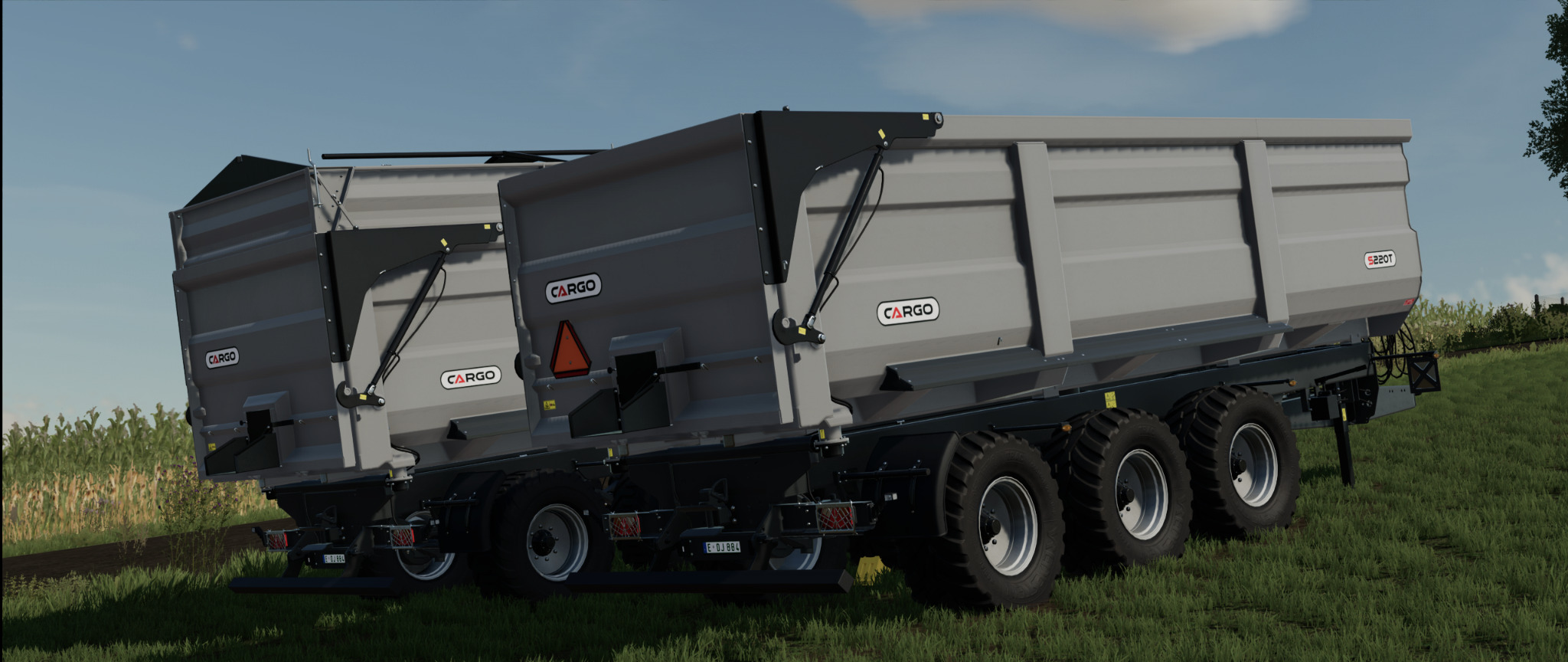 Dwie szare przyczepy rolnicze Cargo S220T stoją na polu z zieloną trawą i kukurydzą w tle pod błękitnym niebem.