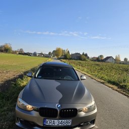 Patryk Kmieć - Szary samochód BMW z czarnym dachem panoramicznym zaparkowany na wąskiej asfaltowej drodze wiejskiej w słoneczny dzień, widok z przodu na tle pól i zabudowań.