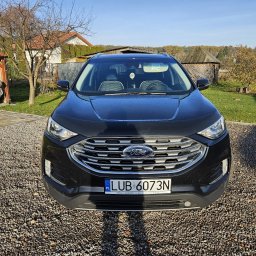 Patryk Kmieć - Czarny Ford Edge, widok z przodu, na żwirowym podjeździe, z delikatnie przyciemnioną szybą przednią, widoczne drzewa i budynki w tle.