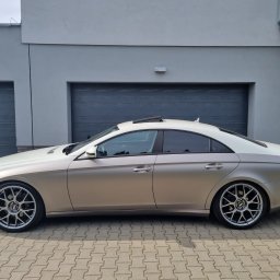 Patryk Kmieć - Mercedes CLS w złotym oklejeniu, z przyciemnionymi szybami, zaparkowany na kostce brukowej przed szarym budynkiem z dwoma garażami.