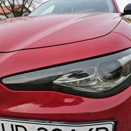 Patryk Kmieć - Czerwony samochód marki Alfa Romeo, widok z przodu, z widoczną przyciemnioną przednią lampą, zarys drzew na masce samochodu