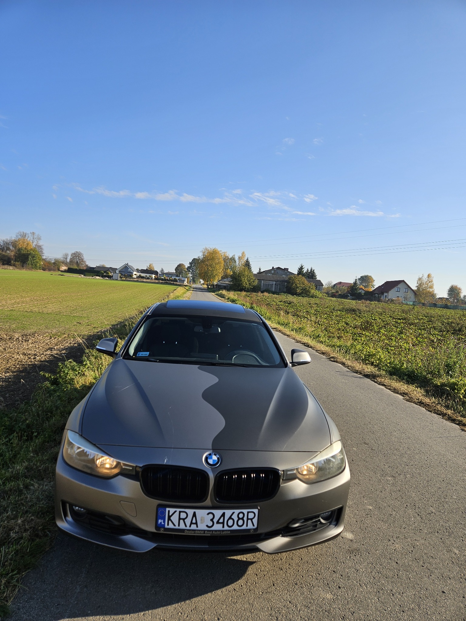 Szary samochód BMW z czarnym dachem panoramicznym zaparkowany na wąskiej asfaltowej drodze wiejskiej w słoneczny dzień, widok z przodu na tle pól i zabudowań.