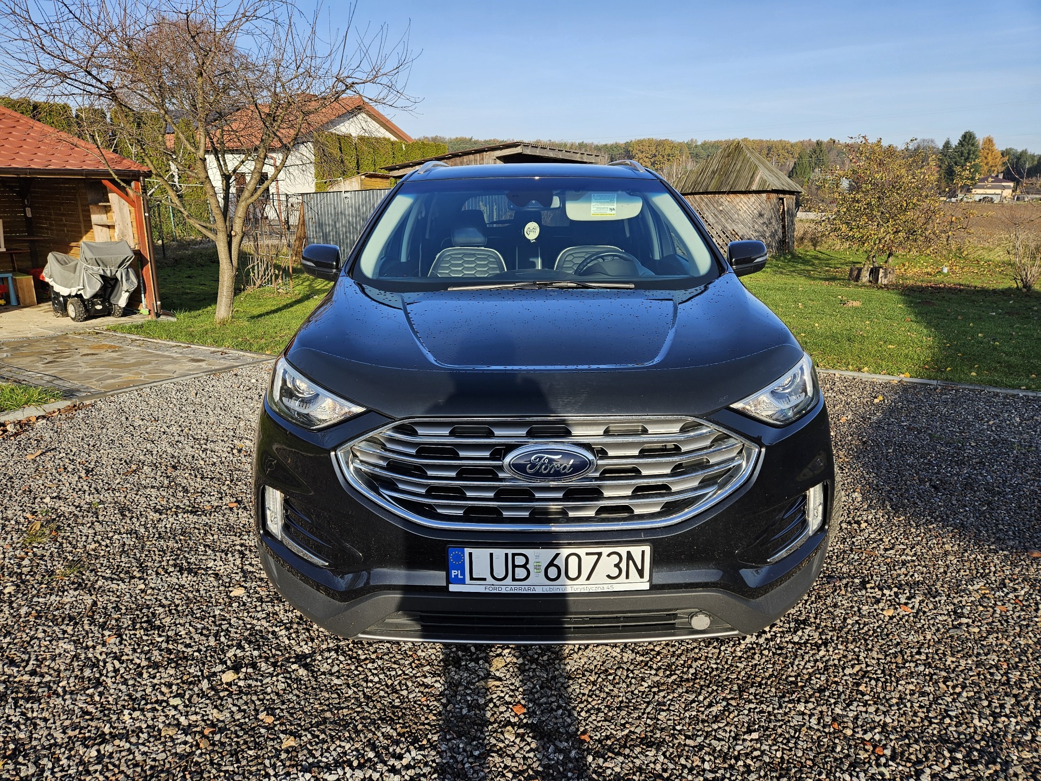 Czarny Ford Edge, widok z przodu, na żwirowym podjeździe, z delikatnie przyciemnioną szybą przednią, widoczne drzewa i budynki w tle.