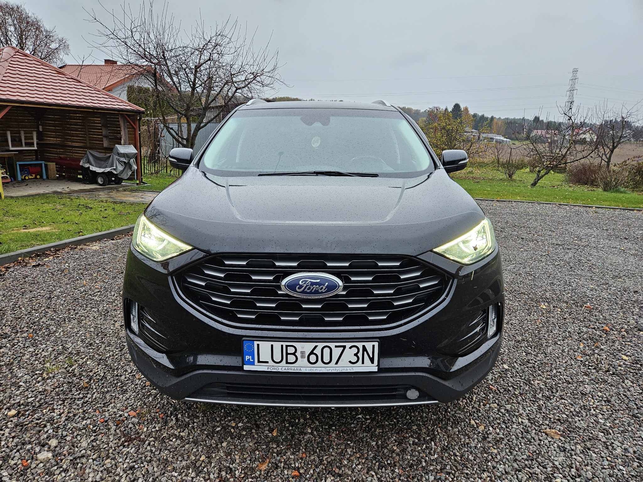 Czarny Ford Edge zaparkowany na żwirowym podjeździe, widok z przodu, reflektory włączone, tablica rejestracyjna z Lublina.