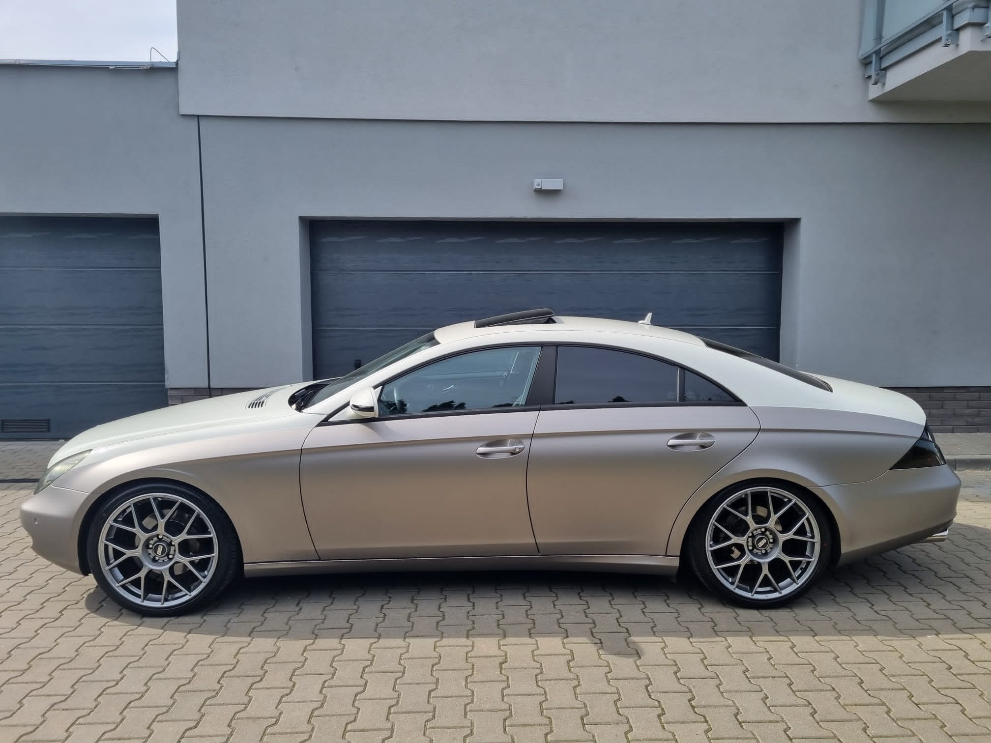 Mercedes CLS w złotym oklejeniu, z przyciemnionymi szybami, zaparkowany na kostce brukowej przed szarym budynkiem z dwoma garażami.