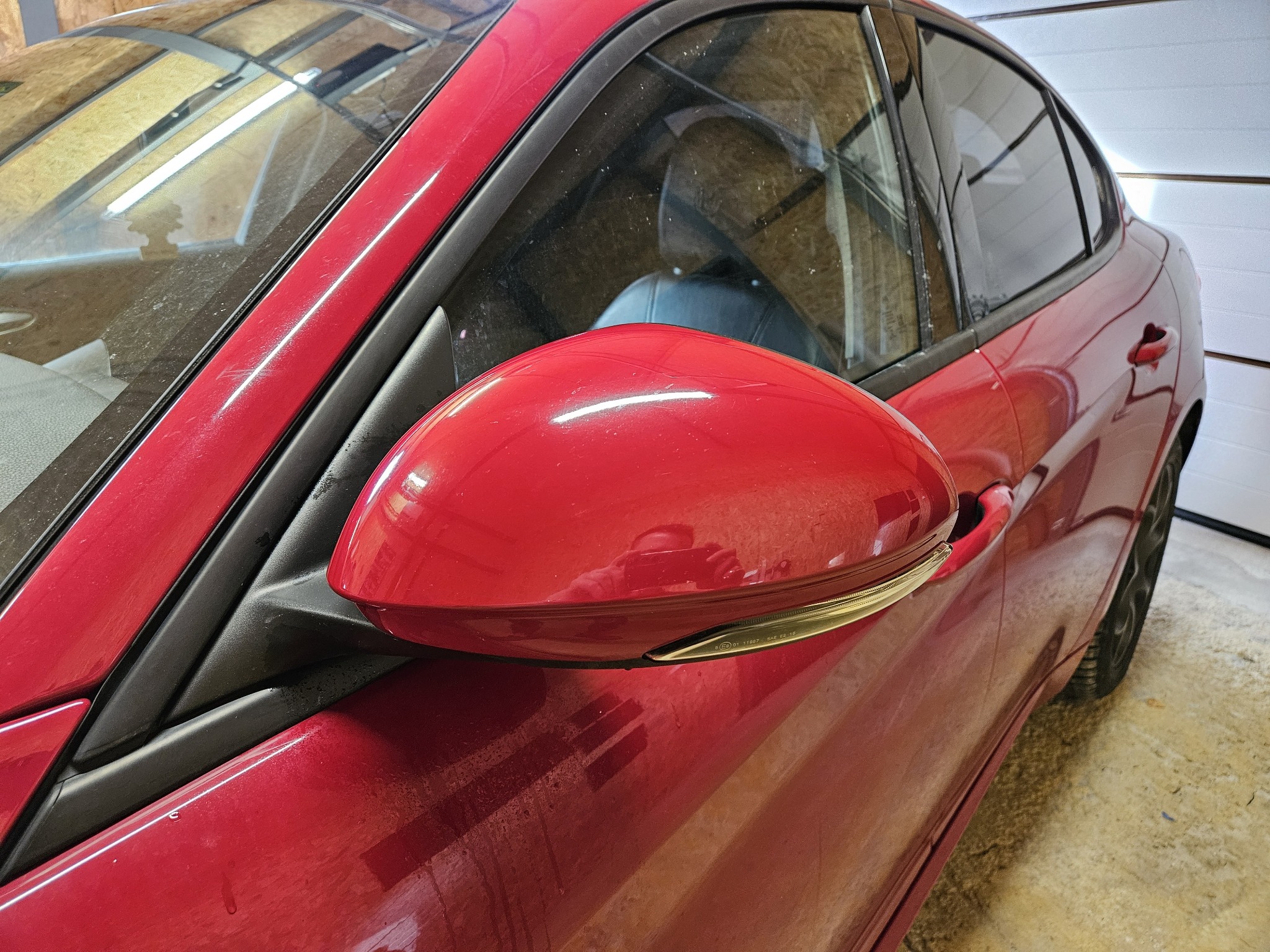 Czerwony samochód z widocznym, błyszczącym lusterkiem bocznym, lekko zabrudzone szyby i karoseria, zarys wnętrza auta.