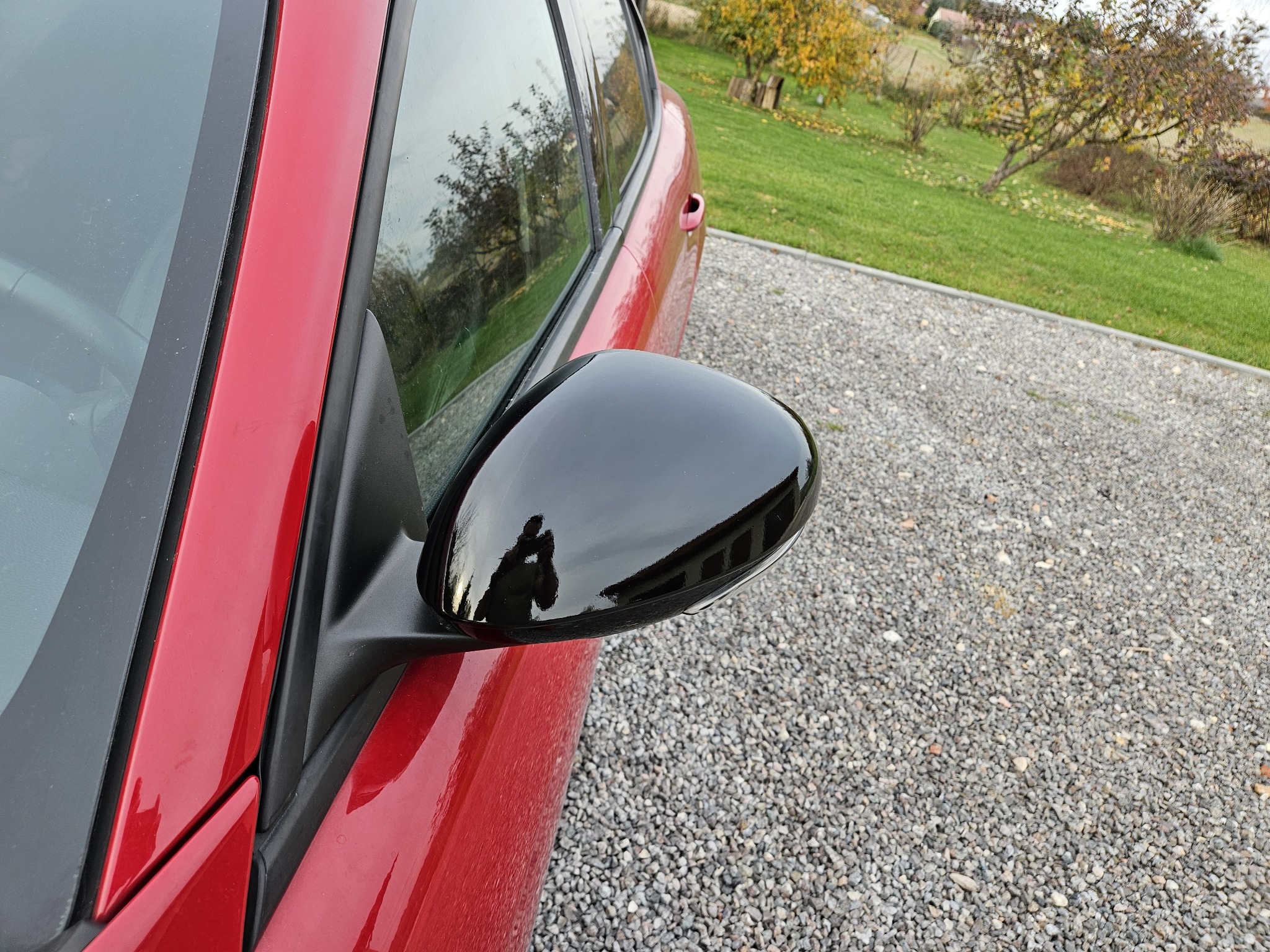 Błyszczący, czarny, lakierowany lusterko boczne na czerwonym samochodzie, z odbiciem osoby robiącej zdjęcie, na tle żwirowego podjazdu i zielonego trawnika z drzewami.