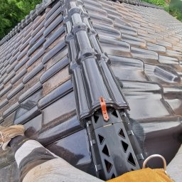 Stanbud - Montaż czarnej dachówki ceramicznej z elementami wentylacyjnymi, widok z perspektywy dekarza w roboczych butach i spodniach.