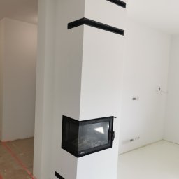 Stanbud - Biały, nowoczesny kominek narożny z czarnymi akcentami w nowo wykończonym wnętrzu, widoczne zabezpieczenia podłogi na czas prac wykończeniowych.