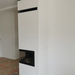 Stanbud - Nowoczesny kominek w obudowie z białego gładkiego tynku z czarnymi poziomymi wstawkami, w trakcie wykańczania, widoczna surowa podłoga zabezpieczona taśmą.