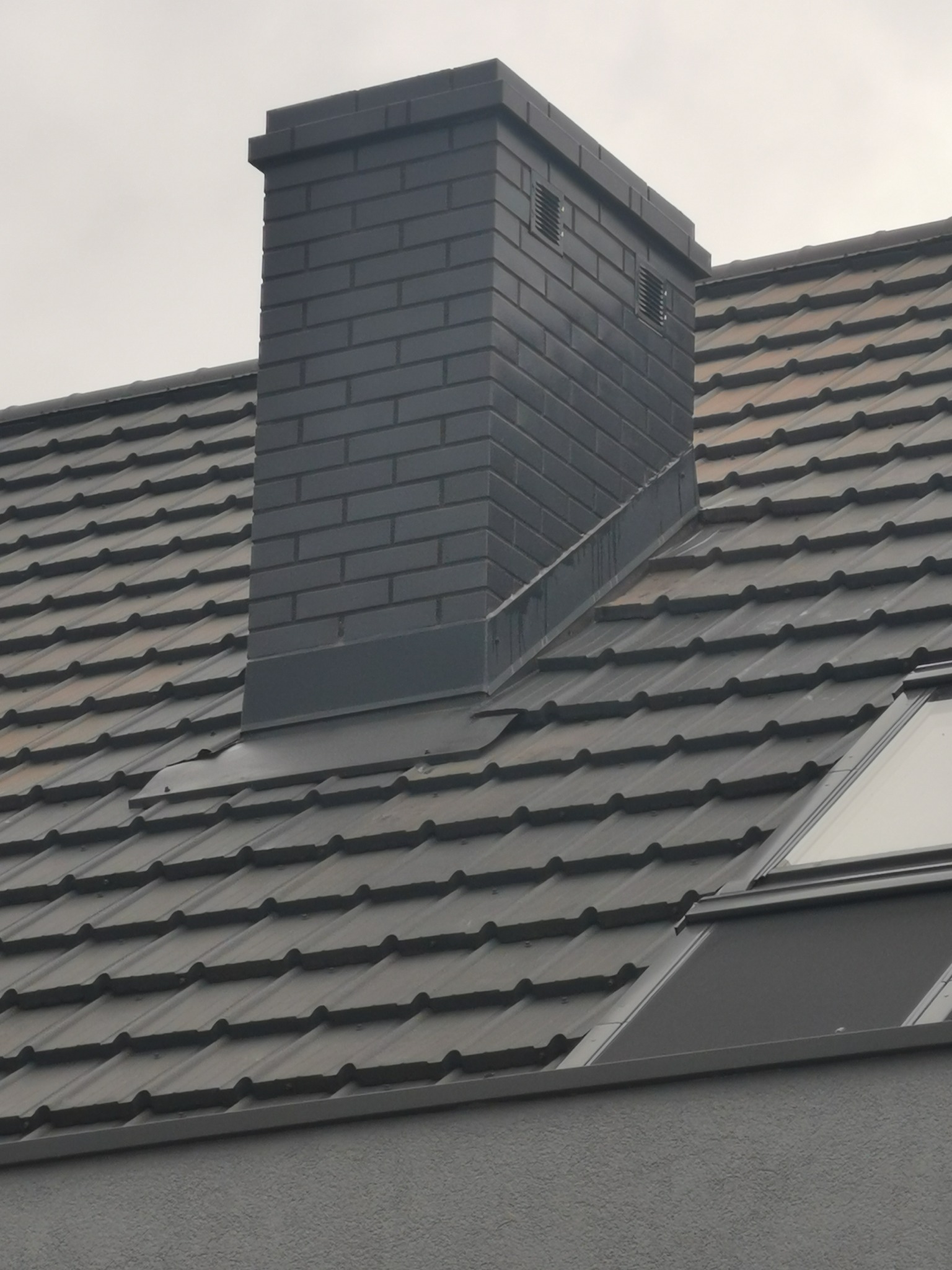 Grafitowy komin z cegły klinkierowej z kratkami wentylacyjnymi, wystający ponad dachówkę ceramiczną w kolorze antracytowym, na tle pochmurnego nieba.